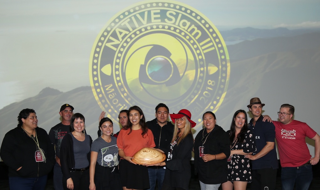 The NATIVE Slam III filmmakers stand in front of the NATIVE Slam logo at Māoriland Film Festival 2018. They are holding a pipi shell carved out of wood and look happy. 