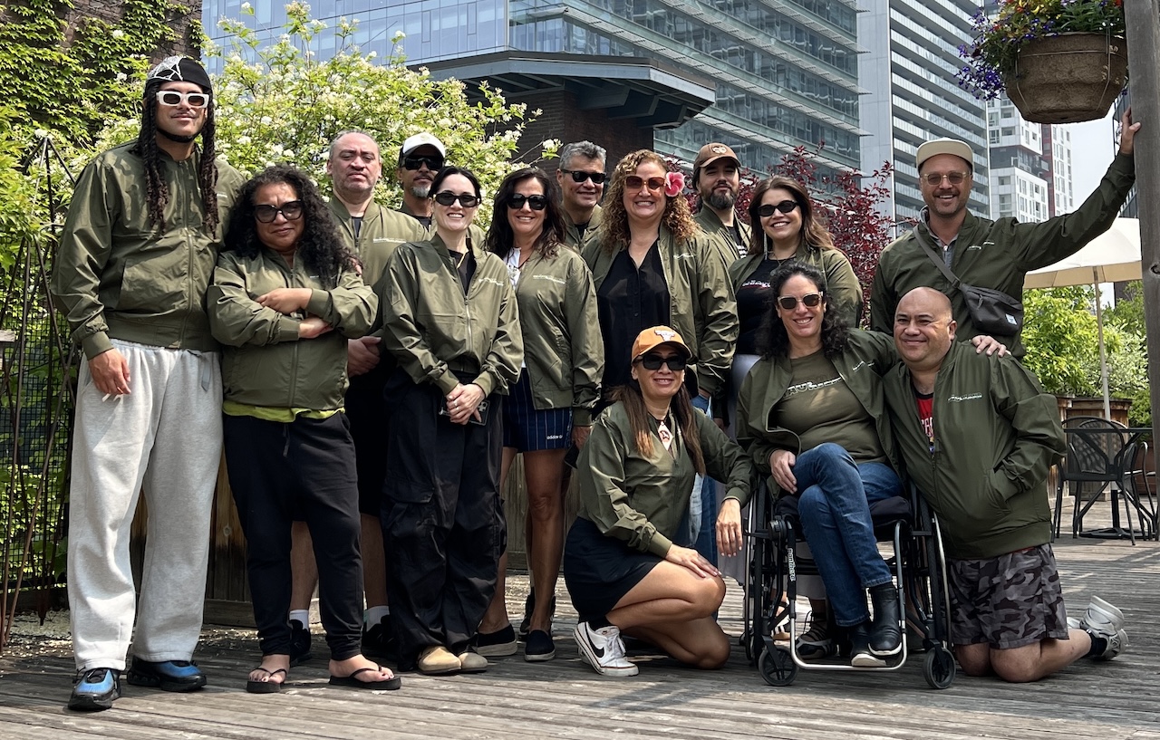The NATIVE Slam III filmmakers stand in front of the NATIVE Slam logo at Māoriland Film Festival 2018. They are holding a pipi shell carved out of wood and look happy. 